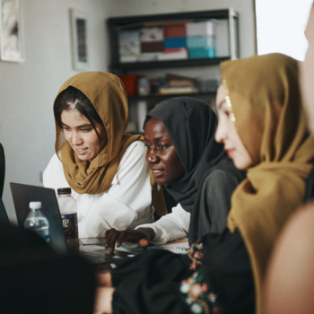 Several women in headscarves lean over a laptop in discussion, seated closely together in an indoor office setting.